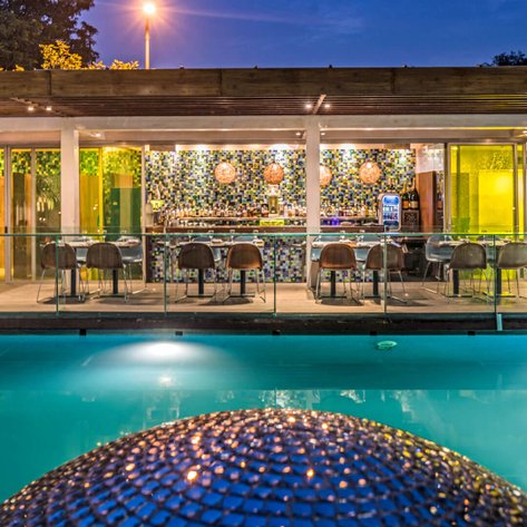 Ein stilvoller Poolbereich mit einer modernen Bar im Hintergrund, beleuchtet von warmem Licht und umgeben von grünen Pflanzen.