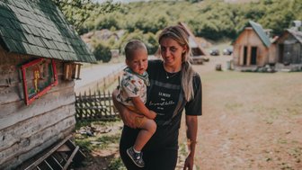 Eine Frau trägt ein kleines Kind in einem malerischen Dorf mit Holzhäusern und grünen Hügeln im Hintergrund.