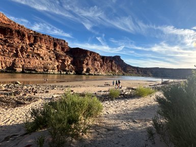 Eine Gruppe von Menschen steht am Ufer des Colorado River, umgeben von beeindruckenden roten Felsen und einem klaren Himmel.