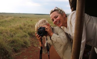 Ein fröhlicher Junge mit einer Kamera posiert, während eine lächelnde Frau ihm zur Seite steht, umgeben von einer weiten Savanne.