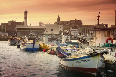Bunte Fischerboote liegen am Hafen, während die Sonne hinter den Gebäuden untergeht und den Himmel in warmen Farben färbt.