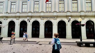 Eine Person mit einem Rucksack geht auf einem gepflasterten Platz vor einem historischen Gebäude mit Flaggen.
