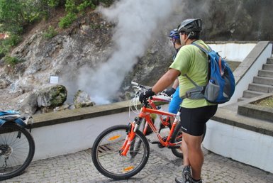 Zwei Mountainbiker stehen an einem rauchenden Felsen, während sie die dampfende Landschaft in Furnas bewundern.