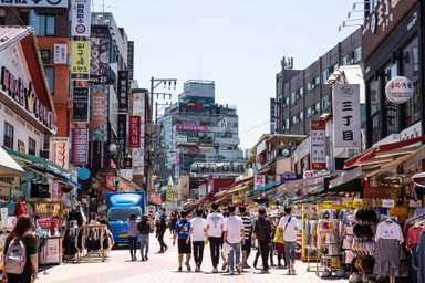 Eine belebte Einkaufsstraße in Hongdae, gesäumt von Geschäften und bunten Schildern, mit vielen Passanten im Sonnenlicht.