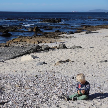 Kind sitzt auf Muscheln am Sandstrand von Boulders Beach – Garden Route Reise mit Kindern