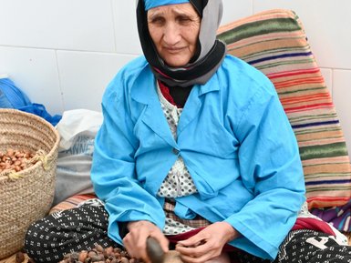 Eine ältere Frau in blauer Kleidung sitzt auf einem Teppich und schält Nüsse, um Arganöl herzustellen.