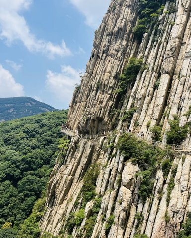 Eine steile Felswand erhebt sich majestätisch über üppige grüne Wälder, während Wanderer auf einem schmalen Pfad entlanggehen.