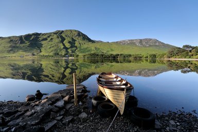 Ein ruhiger See spiegelt die sanften Hügel und den klaren Himmel wider, während ein Boot am Ufer festgemacht ist.