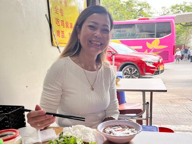 Eine lächelnde Frau sitzt an einem Tisch und genießt eine Mahlzeit mit frischem Salat und Reisnudeln vor sich.