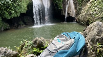Ein blauer Rucksack steht auf einem Felsen, während ein Wasserfall im Hintergrund in einen klaren Teich fließt.