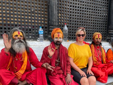 Eine Frau in einem gelben Shirt sitzt lächelnd zwischen vier heiligen Männern in leuchtend roten und orangefarbenen Gewändern.