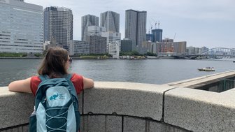 Eine Person mit einem Rucksack blickt über das Wasser auf eine moderne Stadtlandschaft mit hohen Gebäuden.