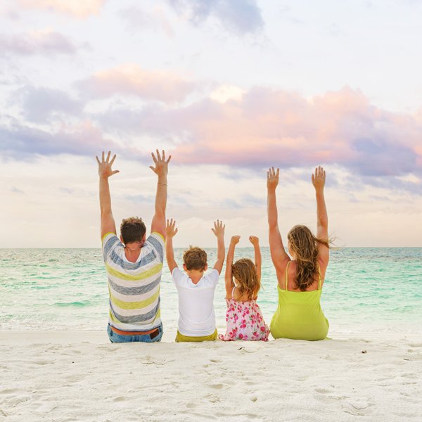 Eine Familie sitzt am Strand, genießt den Sonnenuntergang und hebt die Hände in die Luft, voller Freude und Glück.