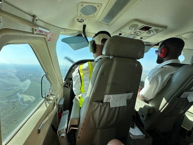 Zwei Piloten sitzen im Cockpit eines Kleinflugzeugs und steuern über eine grüne Landschaft mit Flüssen und Bäumen.