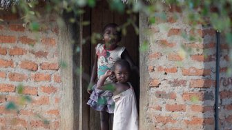 Zwei Kinder stehen in einem offenen Türrahmen eines Lehmhauses, eines lächelt, während das andere überrascht wirkt.