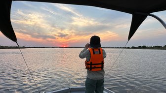 Ein Mensch in einer orangefarbenen Schwimmweste fotografiert den Sonnenuntergang über einem ruhigen Gewässer.