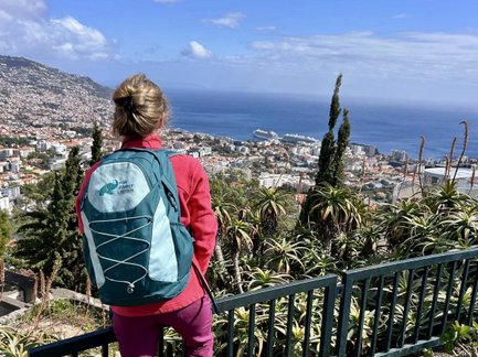 Eine Person mit einem Rucksack steht an einem Geländer und blickt auf die Stadt Funchal und das Meer im Hintergrund.