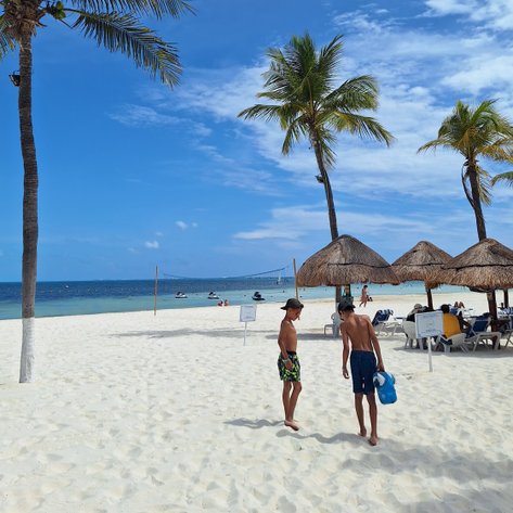 Zwei Jungen gehen barfuß über den weißen Sandstrand, umgeben von Palmen und Sonnenschirmen, während das Meer im Hintergrund glitzert.