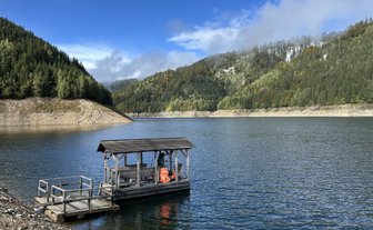 Ein Holzsteg ragt über das ruhige Wasser eines Stausees, umgeben von bewaldeten Hügeln und einem klaren blauen Himmel.