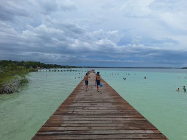 Zwei Kinder laufen auf einem Holzsteg, umgeben von türkisfarbenem Wasser und vielen Schwimmern im Hintergrund.