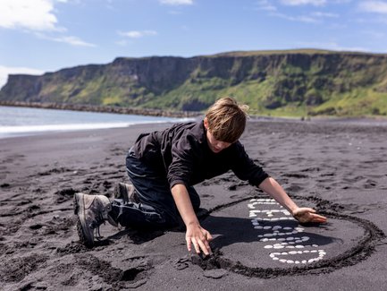 Ein Junge kniet am schwarzen Sandstrand und gestaltet mit Steinen eine kreative Form im Sand.