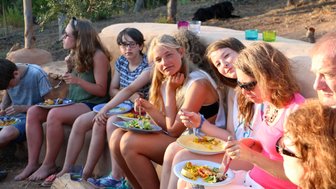 Eine Gruppe von Jugendlichen und Erwachsenen genießt ein gemeinsames Essen im Freien, umgeben von Bäumen und Natur.