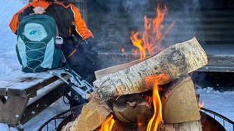 Eine Person in warmer Kleidung sitzt neben einem Lagerfeuer, während Flammen und Rauch in die kalte Luft aufsteigen.