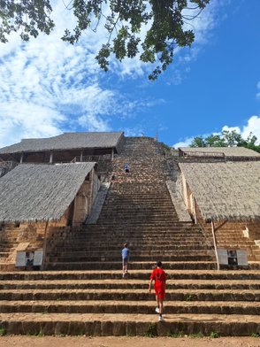 Zwei Kinder steigen die steilen Stufen einer alten Pyramide hinauf, umgeben von grünem Laub und blauem Himmel.