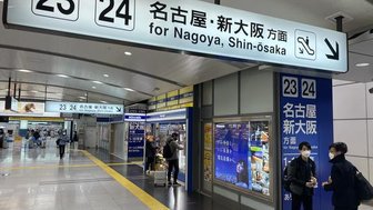 Ein belebter Bahnhof mit Schildern für die Züge nach Nagoya und Shin-Osaka, umgeben von Reisenden und Geschäften.