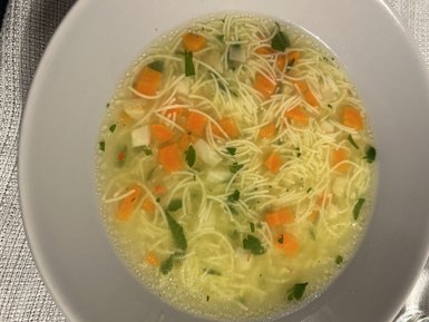 Eine Schüssel mit klarer Brühe, gefüllt mit feinen Nudeln, Karottenstücken und frischen Kräutern, die appetitlich aussieht.