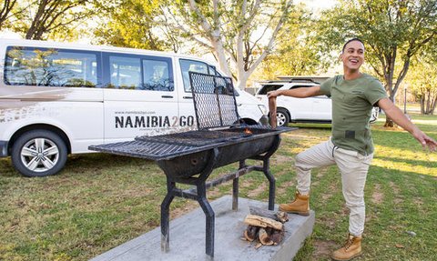 Ein fröhlicher Mann steht neben einem Grill in einem Campingplatz, umgeben von Bäumen und Fahrzeugen im Freien.
