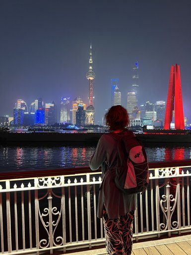 Eine Person mit lockigem Haar steht an einem Geländer und blickt auf die beleuchtete Skyline von Shanghai bei Nacht.