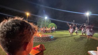 Ein Junge beobachtet eine traditionelle Tanzaufführung am Lagerfeuer, während Zuschauer im Hintergrund sitzen.
