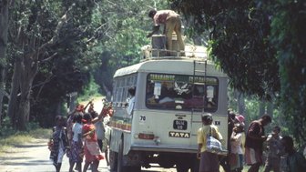 Eine Gruppe von Kindern steht um einen Bus, während ein Mann auf dem Dach Kisten entlädt und die Sonne scheint.