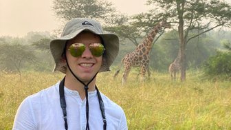 Ein junger Mann mit Sonnenbrille und Hut steht in einer grünen Savanne, während Giraffen im Hintergrund grasen.