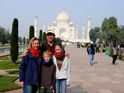 Eine Familie steht lächelnd vor dem Taj Mahal, umgeben von grünen Bäumen und einem klaren Himmel.