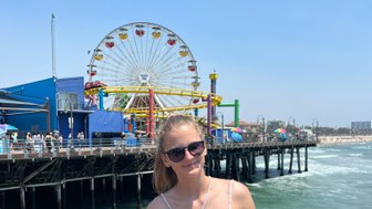 Ein Mädchen steht am Santa Monica Pier, lächelnd, mit einer bunten Achterbahn und dem Ozean im Hintergrund.