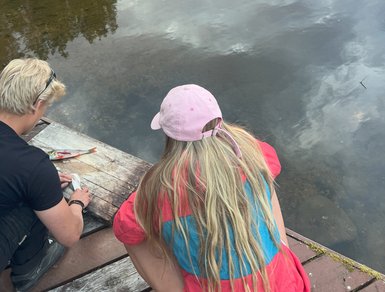Zwei Personen sitzen am Ufer eines ruhigen Sees, während die Sonne hinter den Bäumen untergeht und das Wasser reflektiert.