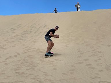 Ein Mann steht auf einem Sandboard und fährt die Düne hinunter, während andere Personen im Hintergrund zuschauen.