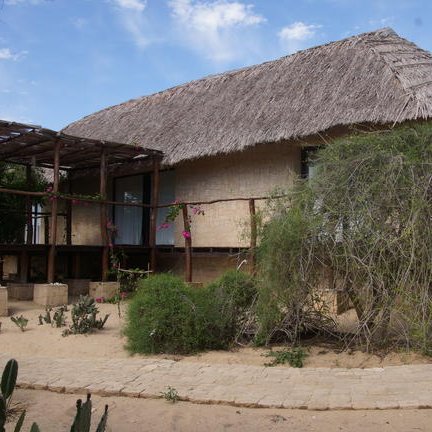 Ein charmantes, strohgedecktes Haus mit Holzveranda, umgeben von üppiger Vegetation und einem sandigen Weg.