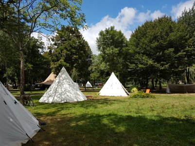 Eine Gruppe von Zelten steht auf einer grünen Wiese, umgeben von Bäumen und einem blauen Himmel mit einigen Wolken.
