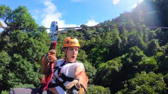 Eine Person mit einem orangefarbenen Helm schwingt fröhlich an einer Zipline über üppige grüne Bäume und strahlend blauem Himmel.