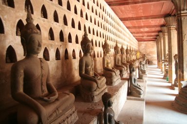 Eine lange Reihe von Buddha-Statuen sitzt in einem historischen Tempel mit kunstvollen Nischen im Hintergrund.
