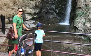 Eine Frau in einem grünen Kleid steht neben einem Kind, das an einem Geländer einen Wasserfall betrachtet.