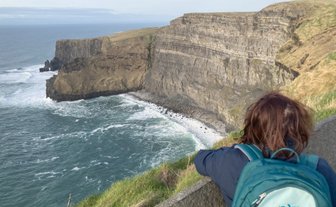 Eine Person mit einem Rucksack steht an der Kante der Klippen und blickt auf die raue See und die zerklüfteten Felsen.