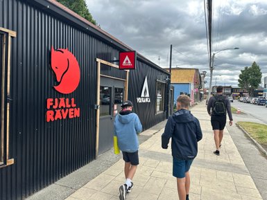 Drei Kinder gehen auf einem Bürgersteig vorbei an Geschäften mit auffälligen Logos in Puerto Natales.
