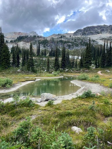Ein ruhiger Teich spiegelt die Wolken und die umliegenden Berge in einer malerischen Landschaft wider.