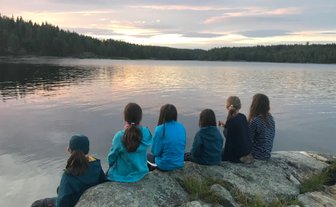 Sechs Kinder sitzen auf einem Felsen am Ufer eines ruhigen Sees und beobachten den Sonnenuntergang über den Bäumen.