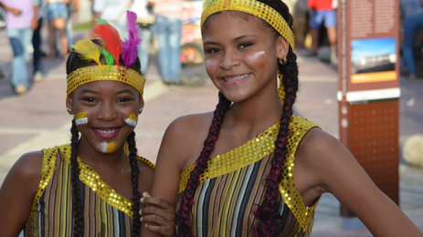 Zwei fröhliche Mädchen in bunten Kostümen lächeln in die Kamera, umgeben von einer lebhaften Menschenmenge.
