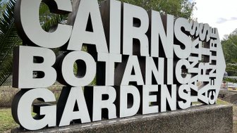 Ein großer, moderner Schriftzug mit den Worten 'Cairns Botanical Gardens' steht im Freien, umgeben von grünen Pflanzen und Bäumen.